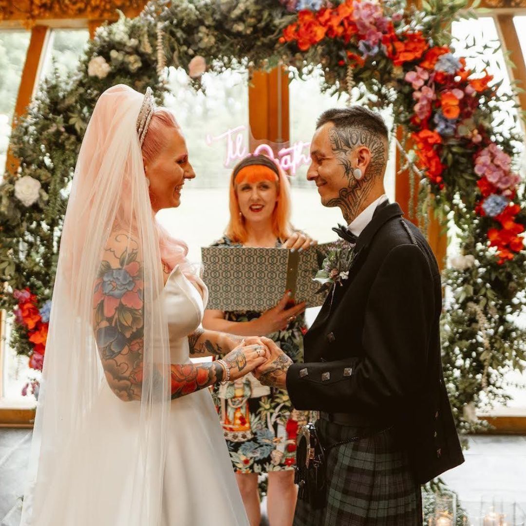 A tattooed couple she as pink hair and a veil and he is wearing a kilt