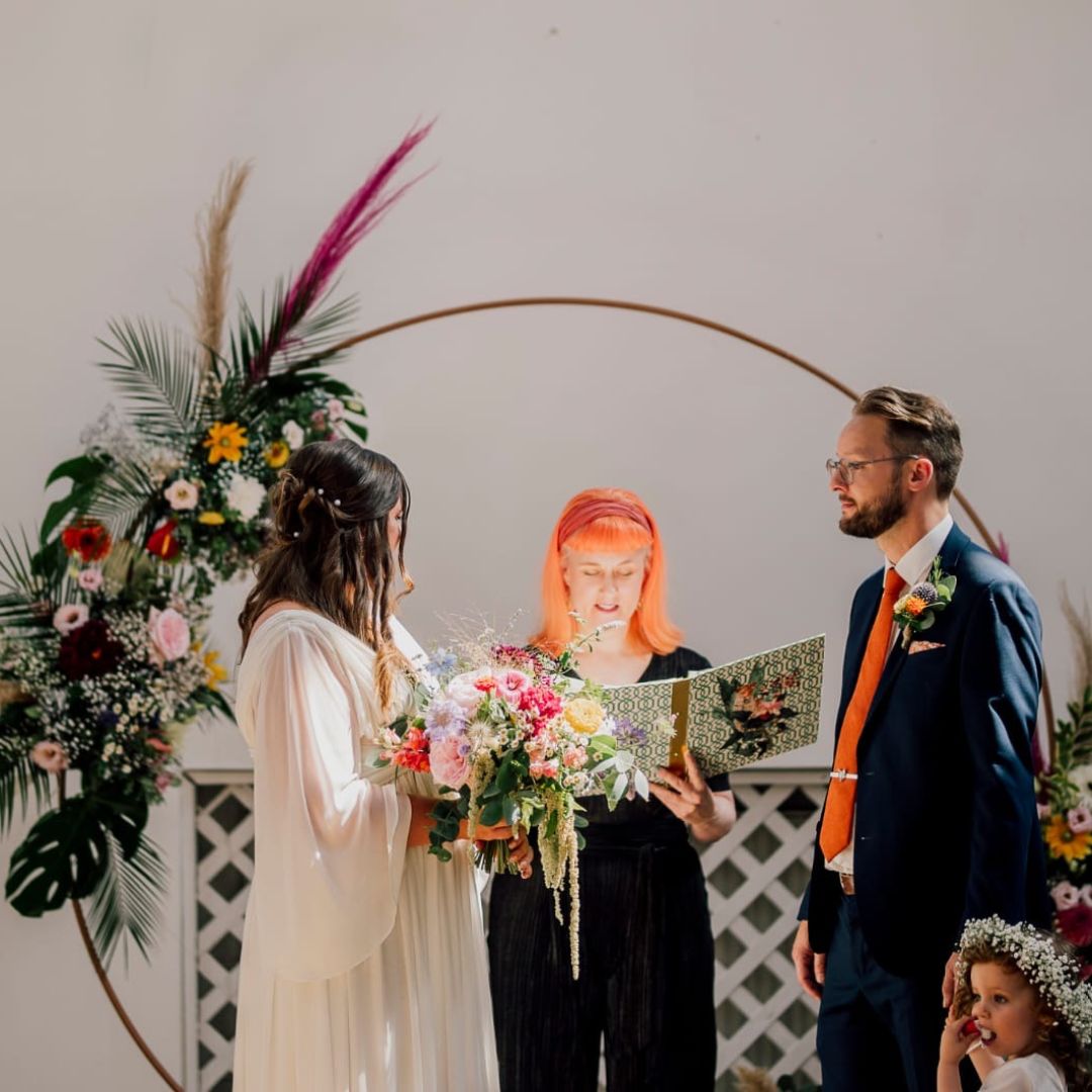 a beautiful photograph of a couple with a circular backdrop with flowers to the left. the light is soft and glowing and catching Jess May's orange hair.