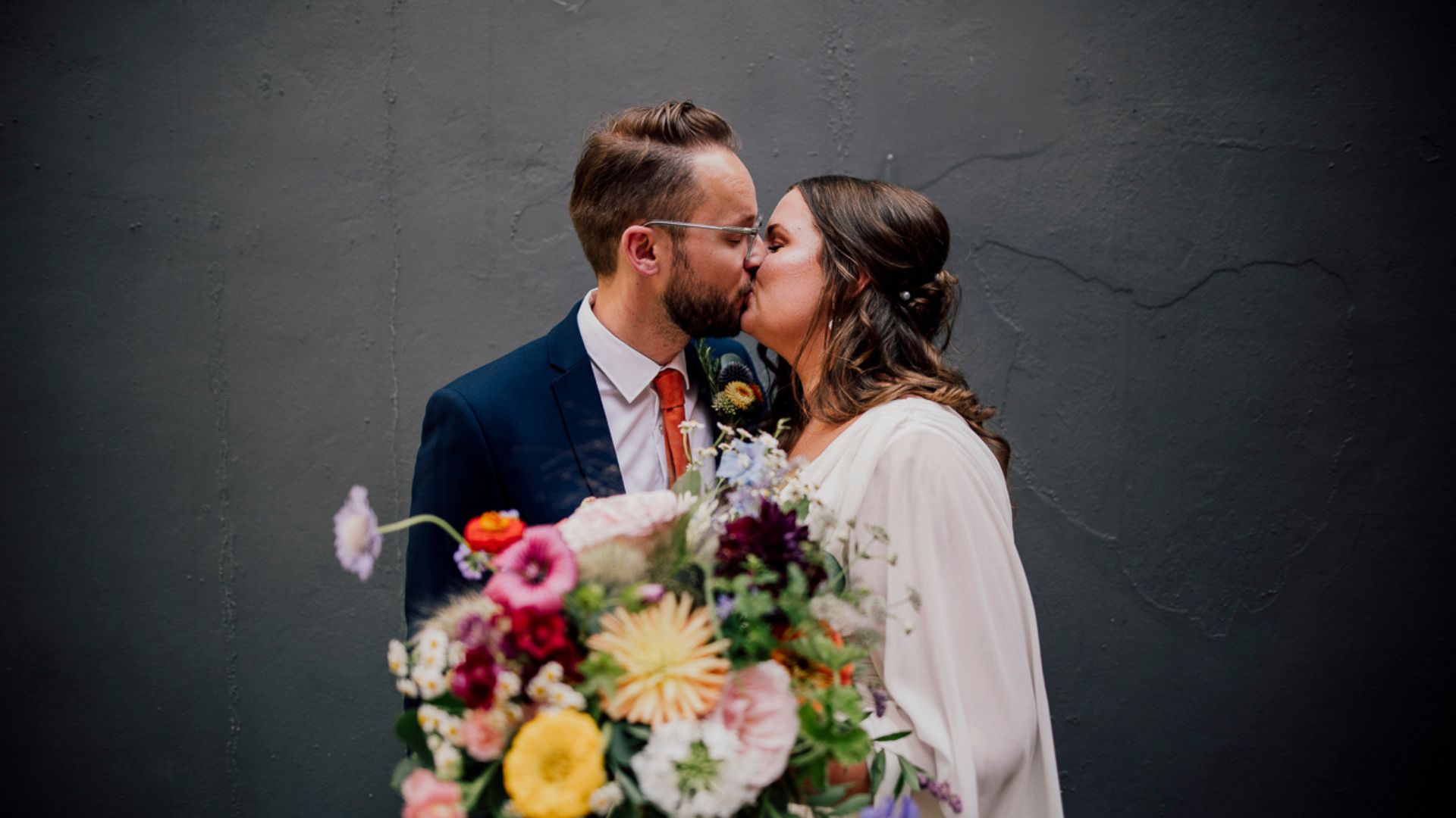 A white heterosexual couple with brown hair and a huge bouquet of flowers in between them and a black backdrop. It is very stylish and she has a white voile dress.