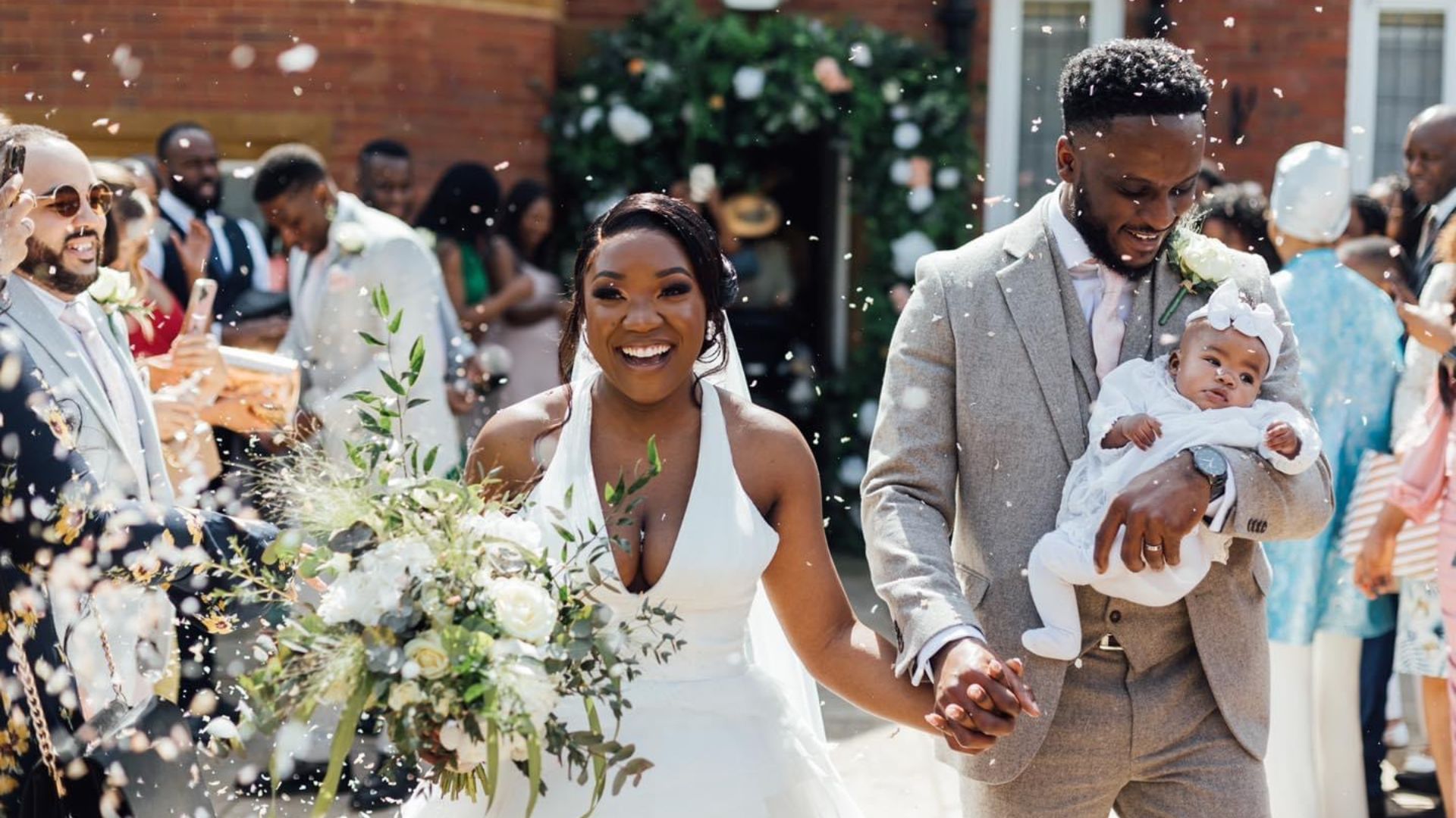 jess may's special days home page (1) A wedding couple smiling and leaving the ceremony. She is wearing a halterneck white dress and has a big bouquet, he is wearing a suit and holding a small baby.