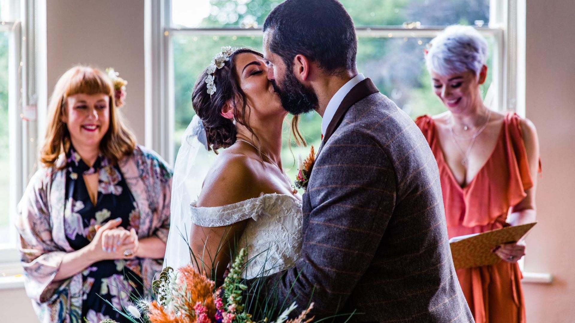 A man and a woman kissing on their wedding day. A woman is reading in the back ground and jess may celebrant is in the left of the image.
