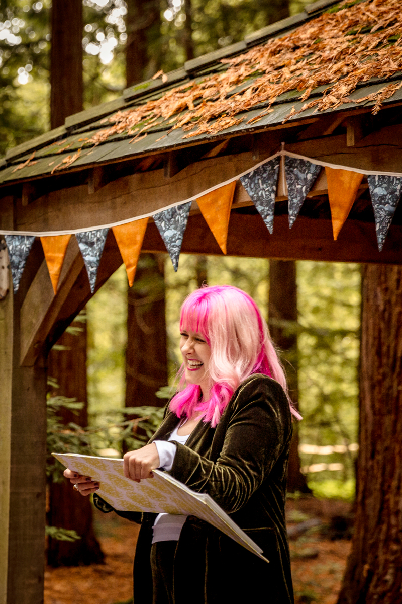 Celebrant-outdoors-wedding Jess May Celebrant laughing and holding her folder in a gazebo in a forest. She is at a wedding in a forest and the gazebo has orange and blue bunting. She is wearing a green velvet suit which is the same colour as the forest behind her. She has pink hair which is vibrant in the green forest.