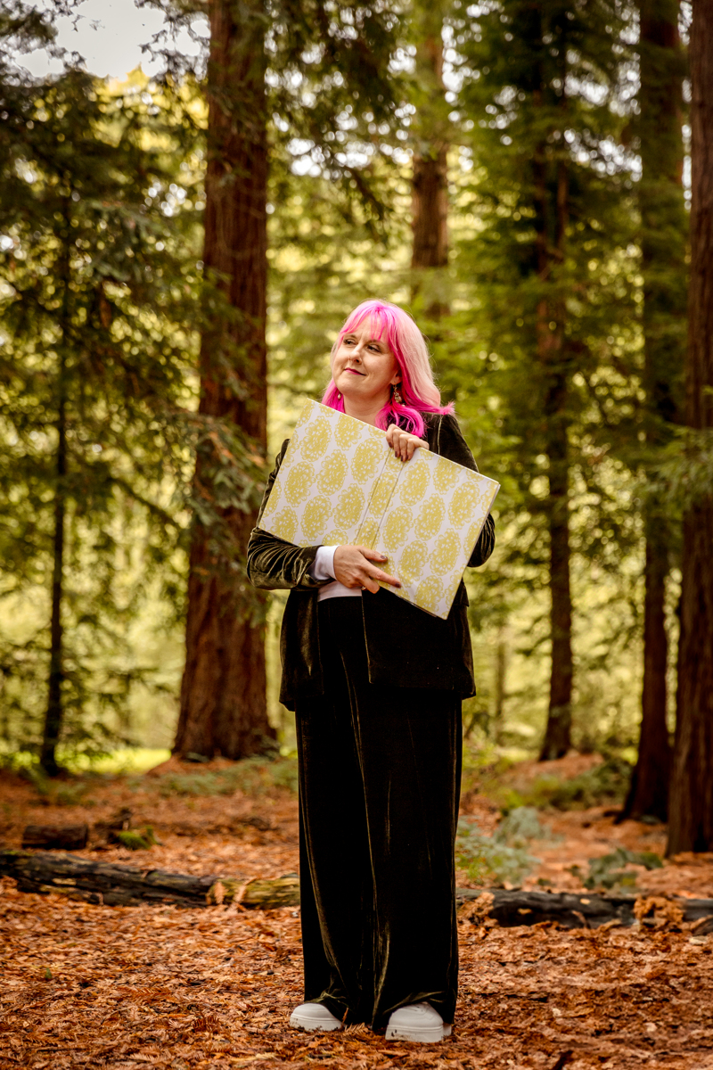 Celebrant-woodland-wedding jess may celebrant with a folder and pink hair and wearing a green velvet suit and gazing into the forest. She is whistful and happy looking on with a look of contentment and satisfaction.