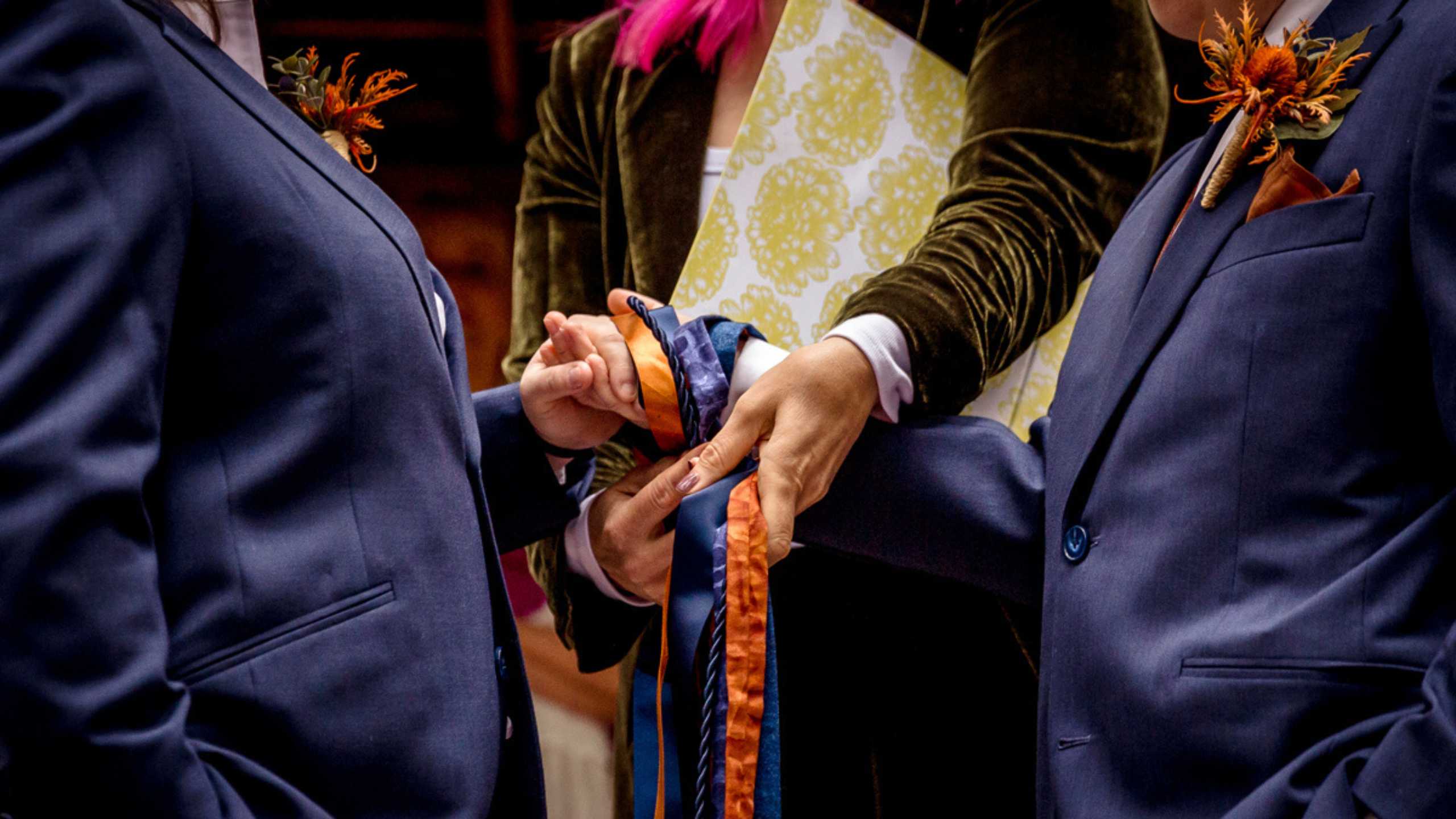 POLYAMOROUS WEDDING CEREMONY two hands being bound together using blue and burnt orange cords. The people are wearing blue jackets.