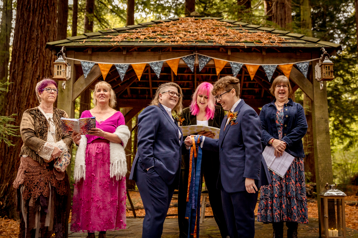 Poly-couple-laughing-wedding A wedding group at the front of the ceremony. Everyone is laughing. There are 6 people. They are all white and different ages. They are under a gazebo in a forest and it has gold and blue bunting. In the centre is a couple with blue suits, one is non-binary. in the centre is a wedding celebrant with pink hair. on either side are the Mother's of the wedding couple, one is smiling and the other looks pensive.