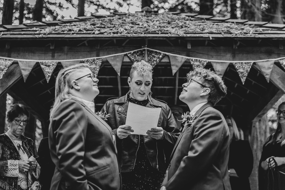 Queer polyamorous woodland wedding ceremony Three people in a black and white photograph taken at a wedding under a gazebo with bunting along the front. Two people are looking at each other and the one in the middle is reading. They are all queer presenting.