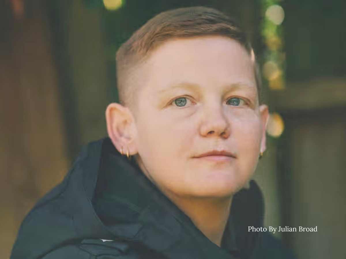 A non-binary person looking thoughtfully at the camera. They have short hair and a masculine presentation. their eyes are green and seem to have pools of water in them. they are wearing small hoop earrings and they are white.