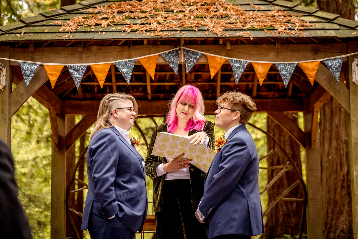 celebrant-with-non-binary-couple Jess May celebrant with pink hair holding a folder in front of a queer couple wearing suits.