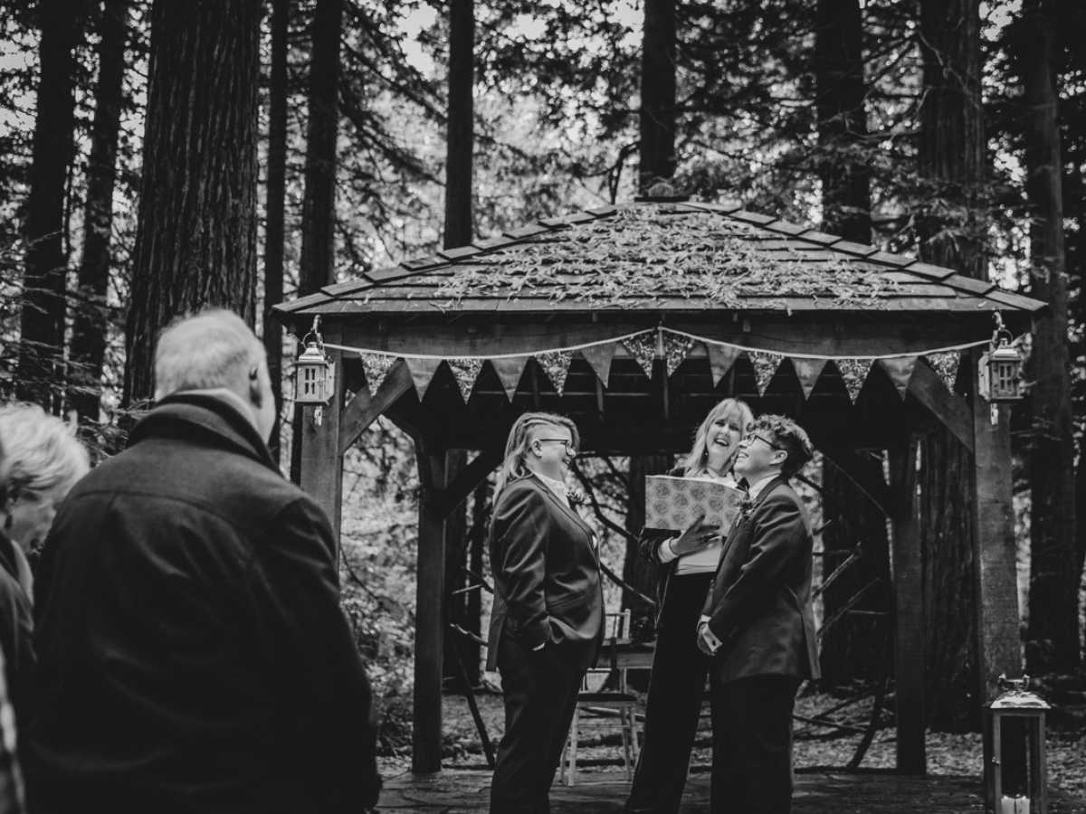 laughter and love at a wedding in a woodland Black and white image of a three people laughing under a gazebo with bunting outside in a forest. Two are getting married and the third is marrying them.