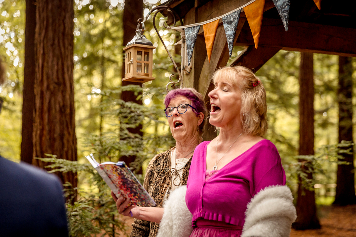 singing in the congregation of a polyamorous wedding Two women in their sixties are singing. One is wearing a bright pink satin dress and has blonde hair and the other has purple hair.