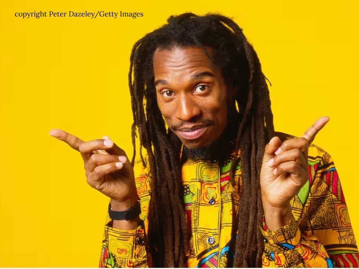Benjamin Zephaniah, a black poet with thread smiling at the camera with an african print shrit and both fingers pointing playfully. The backdrop is a rich bright yellow.