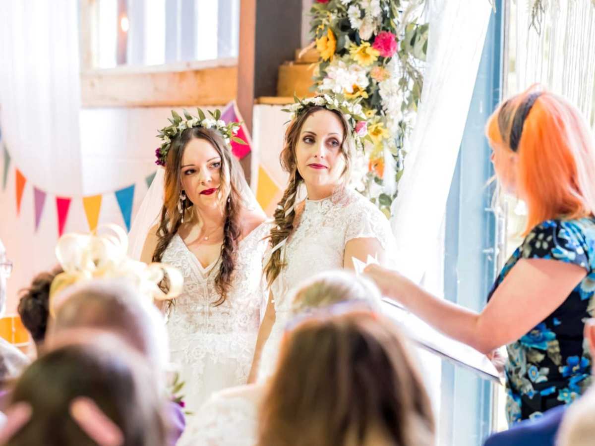 unique wedding readings to reflect your style. two brides dressed in white with dark hair and floral crowns. they are both white and looking thoughtful as a celebrant reads a poem.