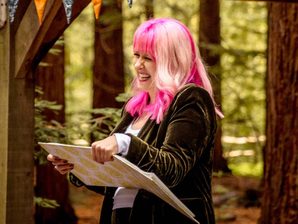unique wedding readings with jess may celebrant Unique Wedding Readings. Jess May Celebrant laughing and holding her folder in a gazebo in a forest. She is at a wedding in a forest and the gazebo has orange and blue bunting. She is wearing a green velvet suit which is the same colour as the forest behind her. She has pink hair which is vibrant in the green forest.