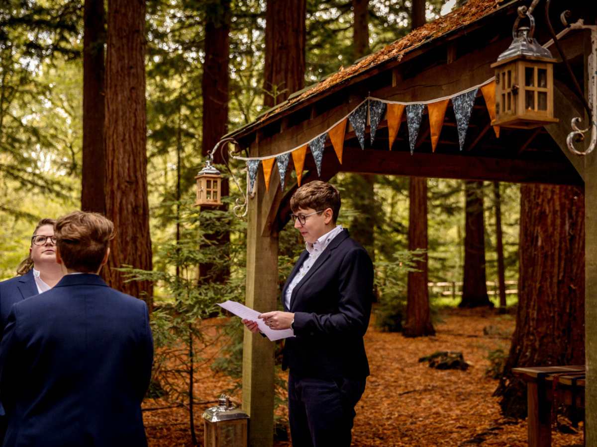 wedding for polyamorous (1) A person in a blue suit holding a piece of paper and reading is standing under a gazebo in an autumnal light. There is bunting and a lantern. Two people are standing to the front of this person.