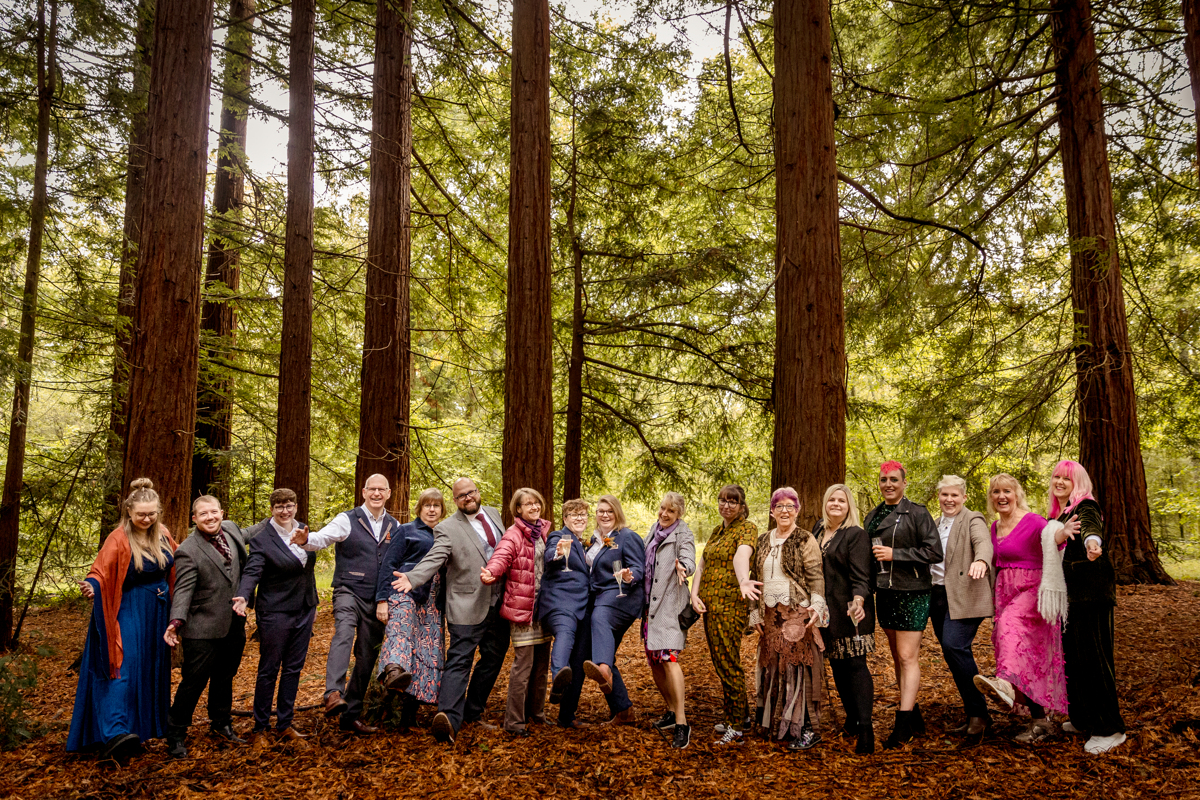 wedding-party-redwood-forest A beautiful happy scene in nature at a woodland wedding. The whole wedding party is assembled in a line in a redwood forest for a Queer Polyamorous couple. People are wearing lots of bright colours and look dwarfed by the enormous redwood trees. Everyone is being playful for the camera.