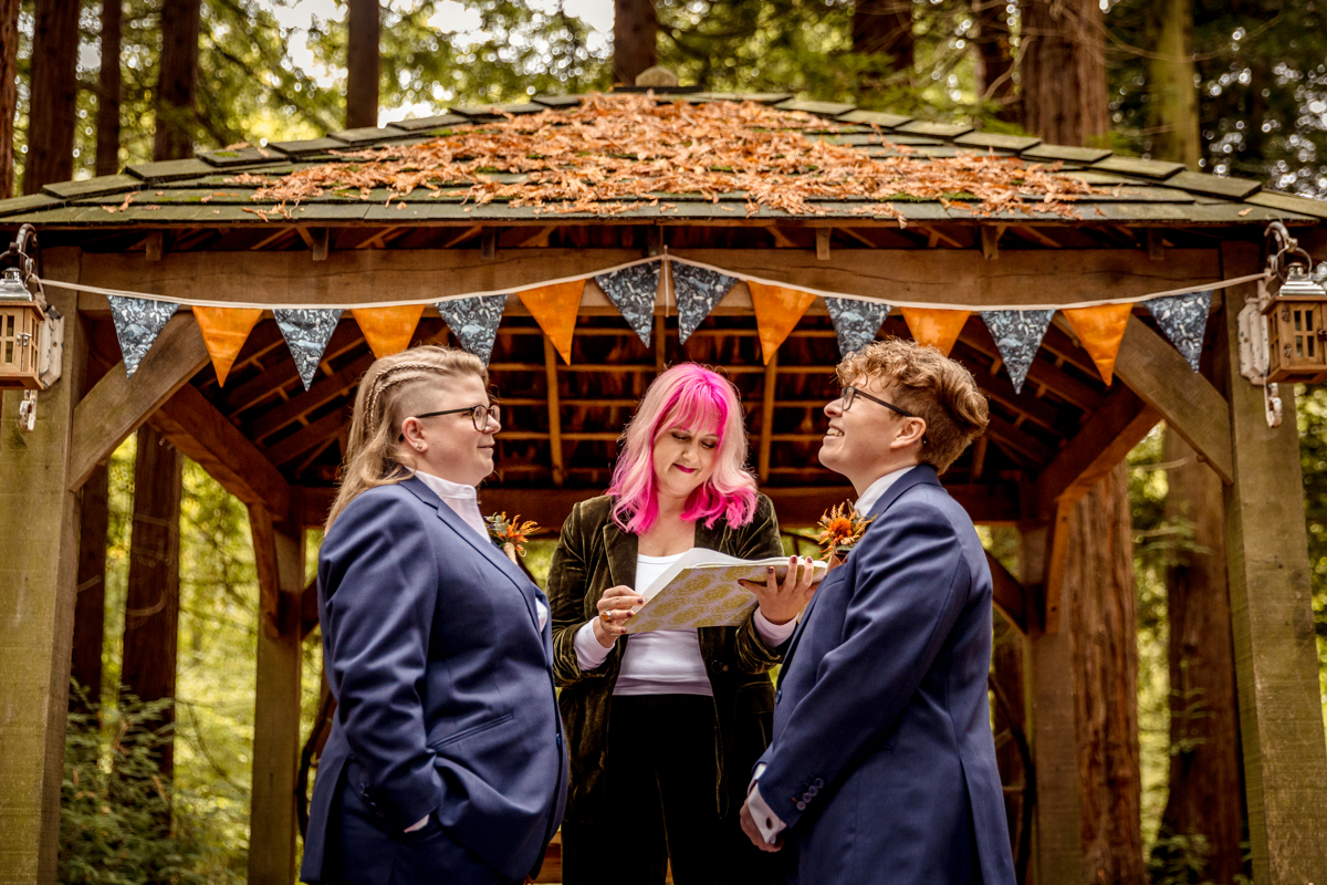 Polyamorous wedding ceremony story woodland poly wedding Polyamorous wedding ceremony in a woodland setting. Two people wearing dark blue suits are stood opposite each other. Both are white and their gender is unclear. They are laughing. Behind is a gazebo with burnt orange and dark blue bunting. The gazebo has dried leaves as it is Autumn. In between the couple is a woman with pink and blonde hair holding a folder and reading their story.