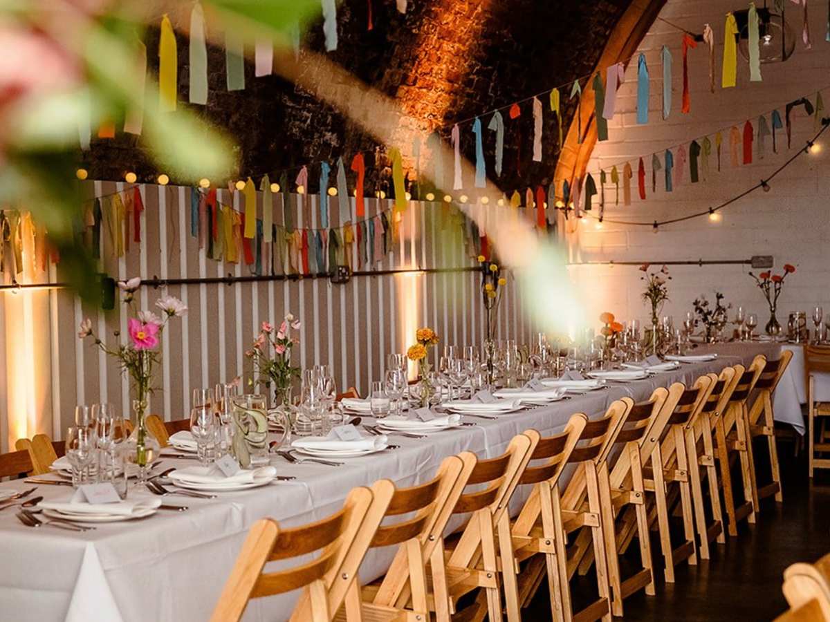 AMP STUDIOS Unusual-London-wedding-venue (1) A cosy space under an archway with a trestle table laid for a wedding. Wooden fold away chairs and bunting in lots of lovely colours
