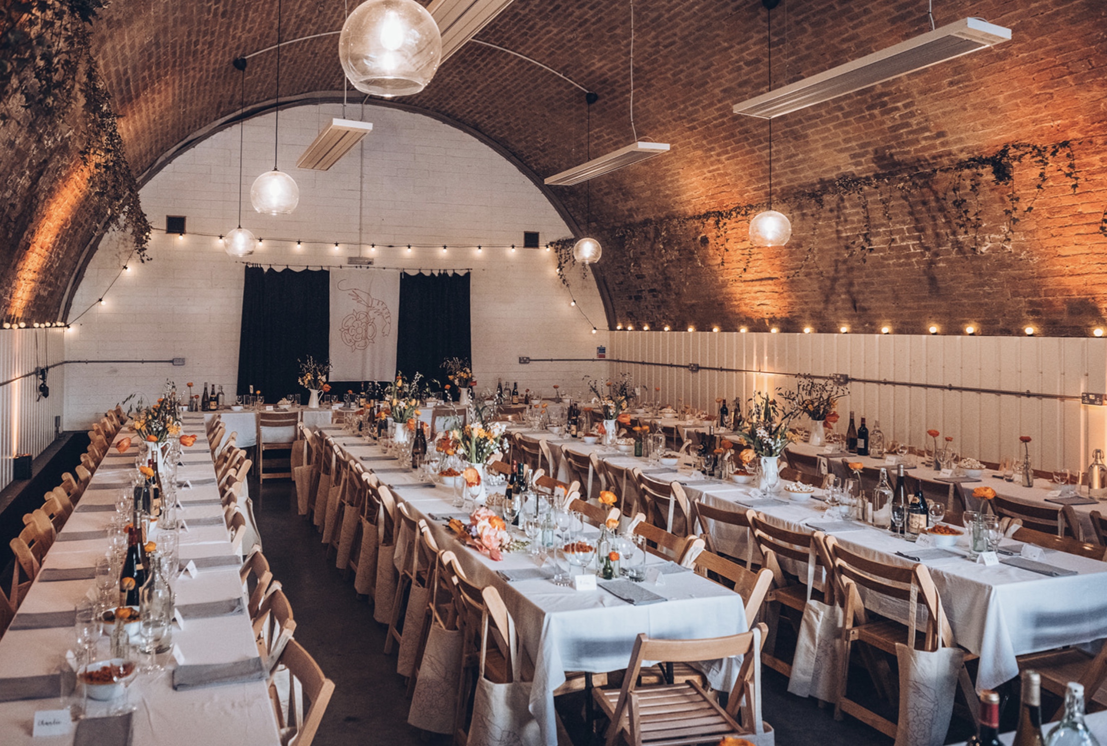 rows of trestle tables laid with table cloths and glasses and flowers for a wedding with wooden picnic chairs under a railway arch strung with fairy lights..