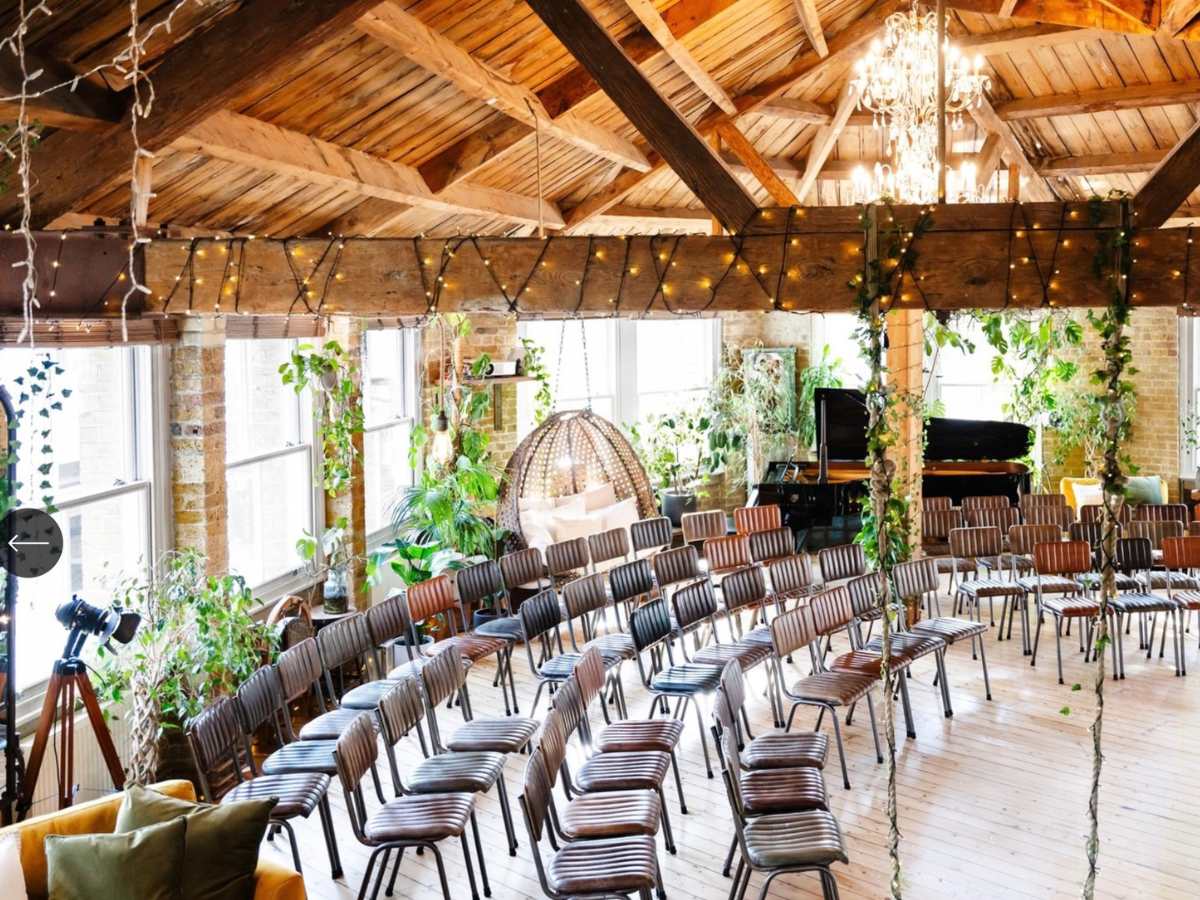 A gorgeous image of a ceremony space set up in a tree house with a swing and curved rows of seats. Plants are everywhere and the light is beautiful. there are fairy lights and in the corner there is a logo for The Shoreditch Tree house