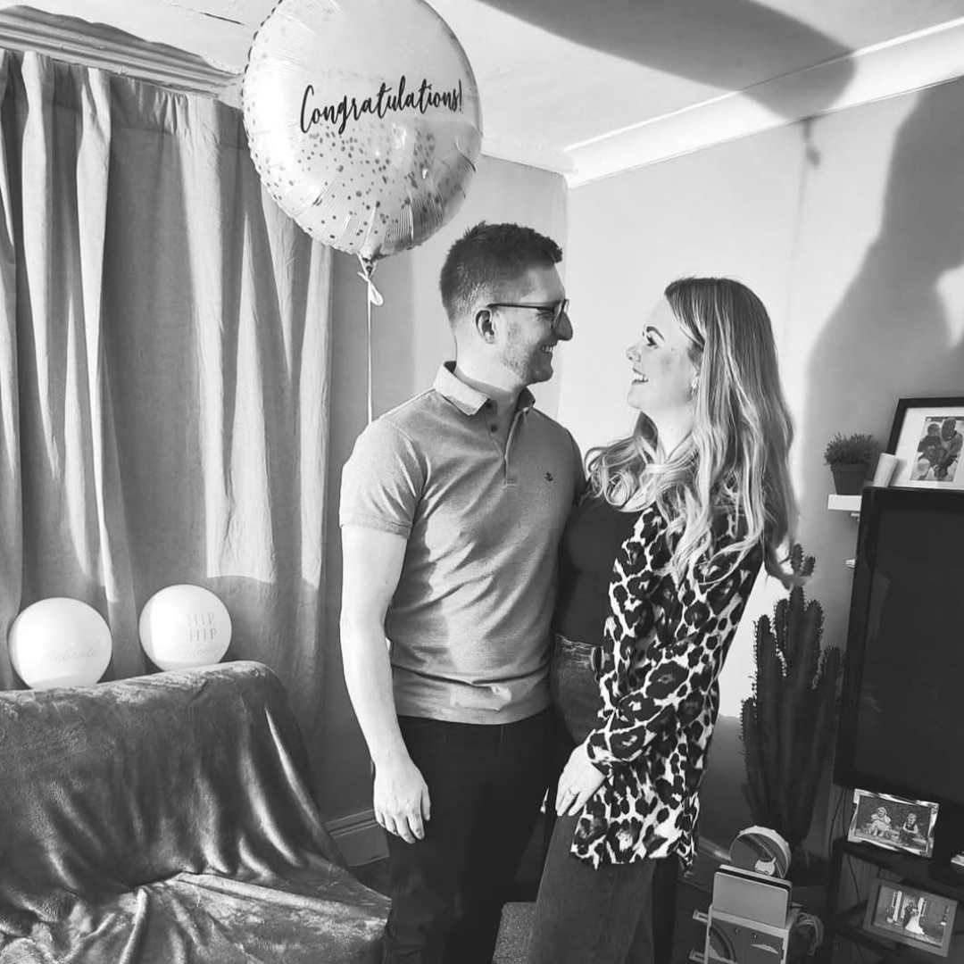 Black & white image of a caucasian couple with a white balloon with congratulations on it. It is an engagment party