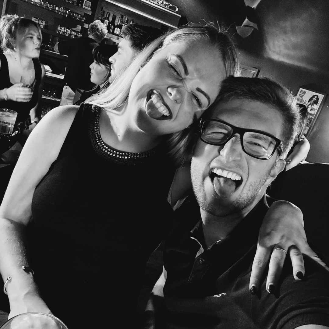 A Black and white photo of a Cis-het couple with their tongues out. She is sitting on his lap. He has glasses on, they are very 'rock n roll'