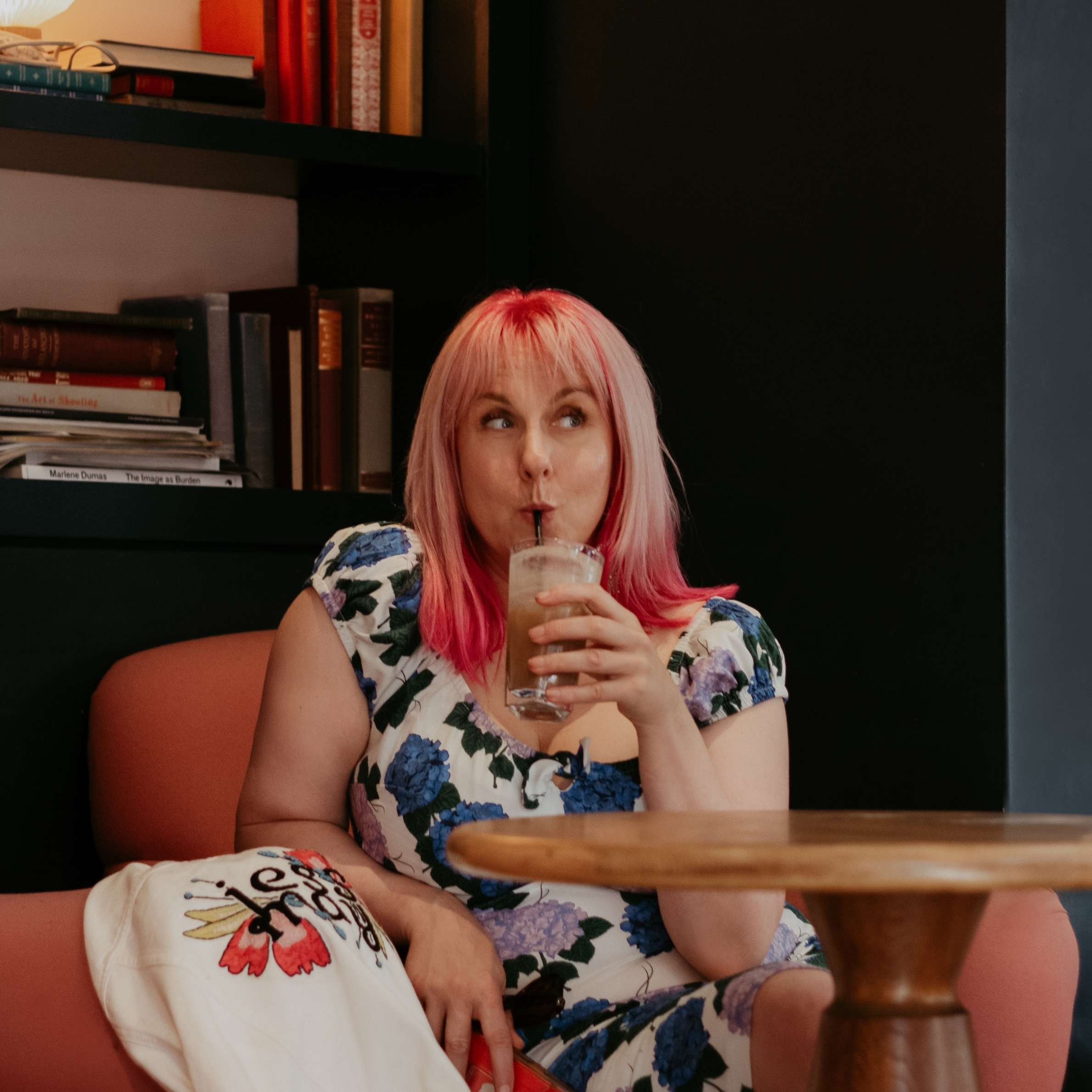 A woman in a blue and white floral dress with pink hair is drinking iced coffee through a straw and looking mischievious.